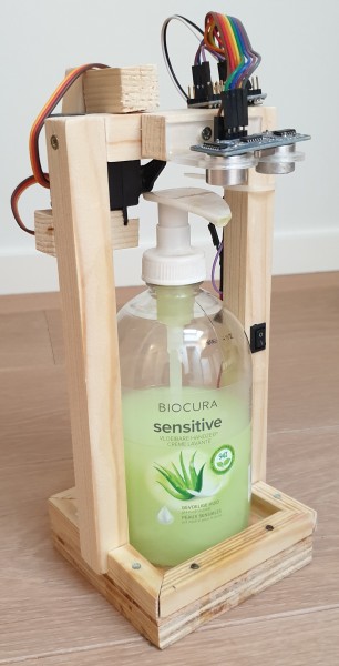 Front view of a wooden construction built around a regular manual soap dispenser bottle. The construction also includes a small-form factor Arduino, a servo, two distance sensors and a lot of wires.